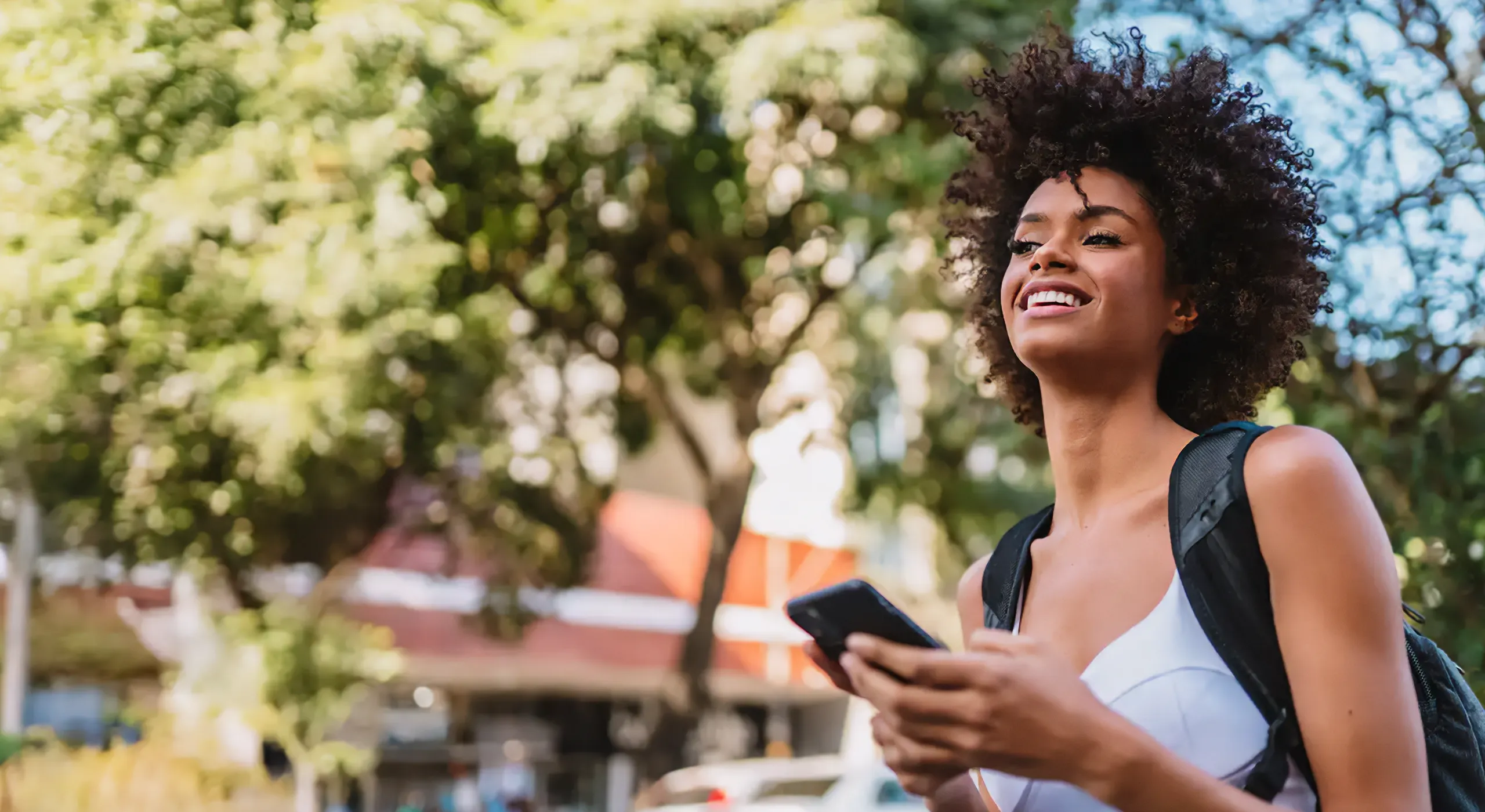young tall woman looking forward and smiling walking downtown with trees in the background on her phone
